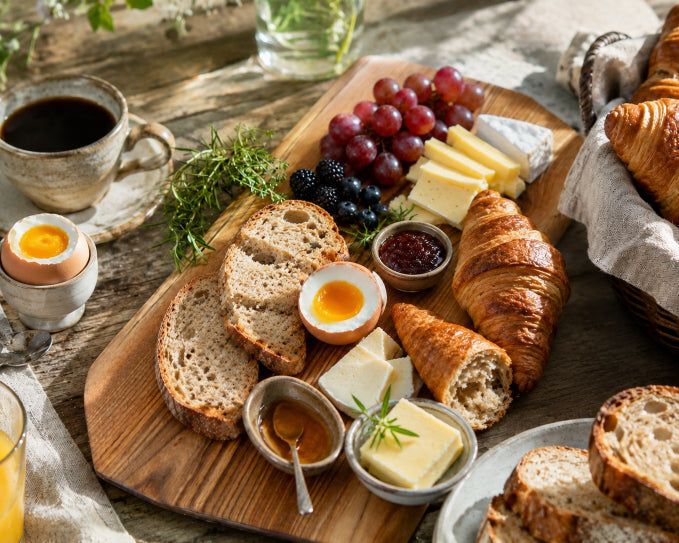     Sonntagsbrunch für sechs Personen mit Croissants, Brot, Käse, Ei, Beeren und Aufstrichen auf massivem Holzbrett