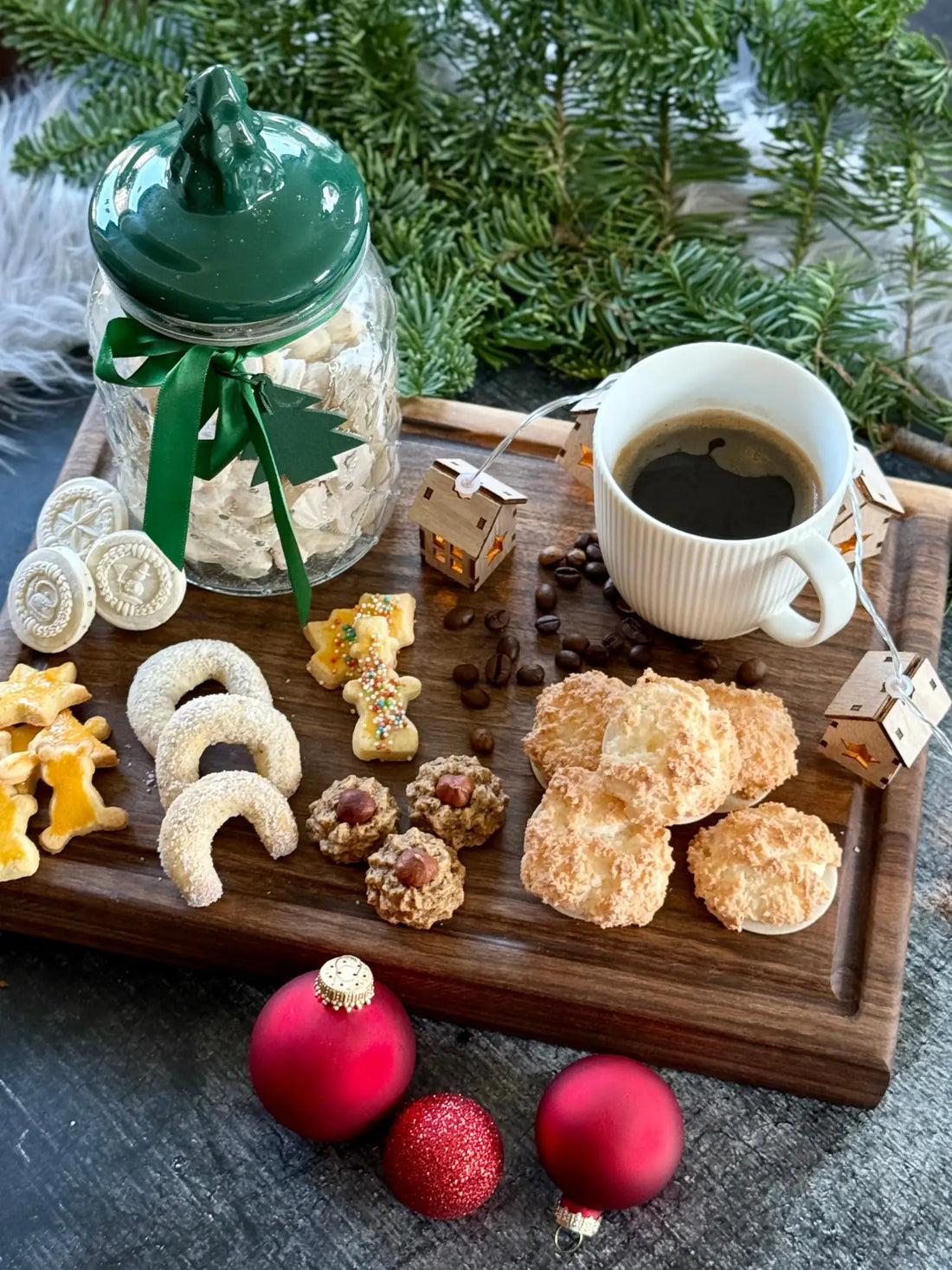 Ein hölzernes Servierbrett mit Weihnachtsplätzchen, Kaffee und roten Ornamenten.