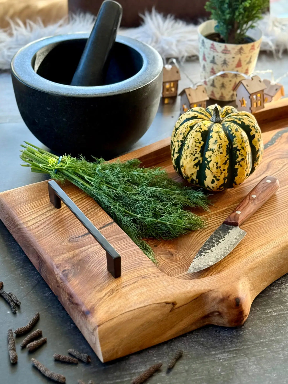 Rustikales Holzschneidebrett mit scharfer Kante, auf dem frischer Dill, ein dekorativer Kürbis und ein schwarzer Mörser mit Stößel zu sehen sind.