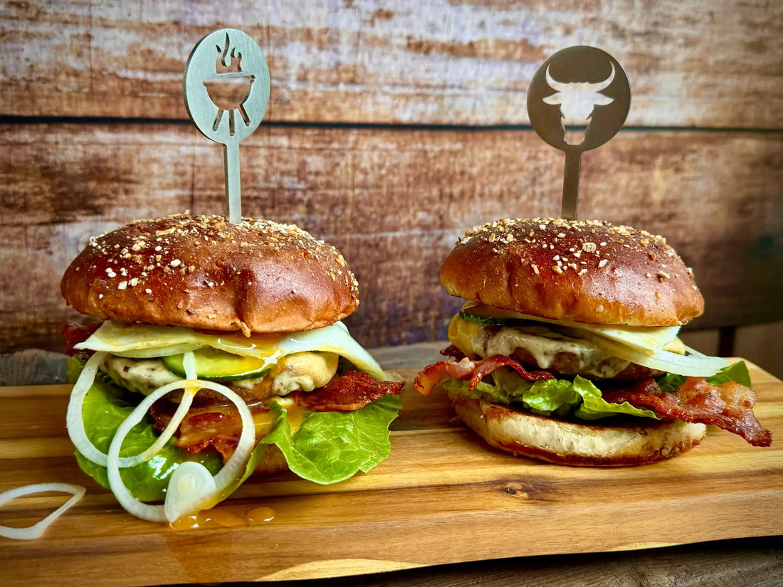 Zwei Gourmet-Burger mit Sesambrötchen, frischem Gemüse und dekorativen Toppings auf einem Holzbrett.